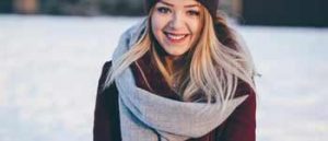 Blonde woman in wine red coat with matching hat and grey scarf with snow in background.