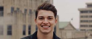 Young brunette man in dark grey jacket in front of blurred tan buildings.
