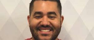 Young Latino man with short cropped hair standing in front of a wall with a diamond pattern in various shades of off-white and light grey. 